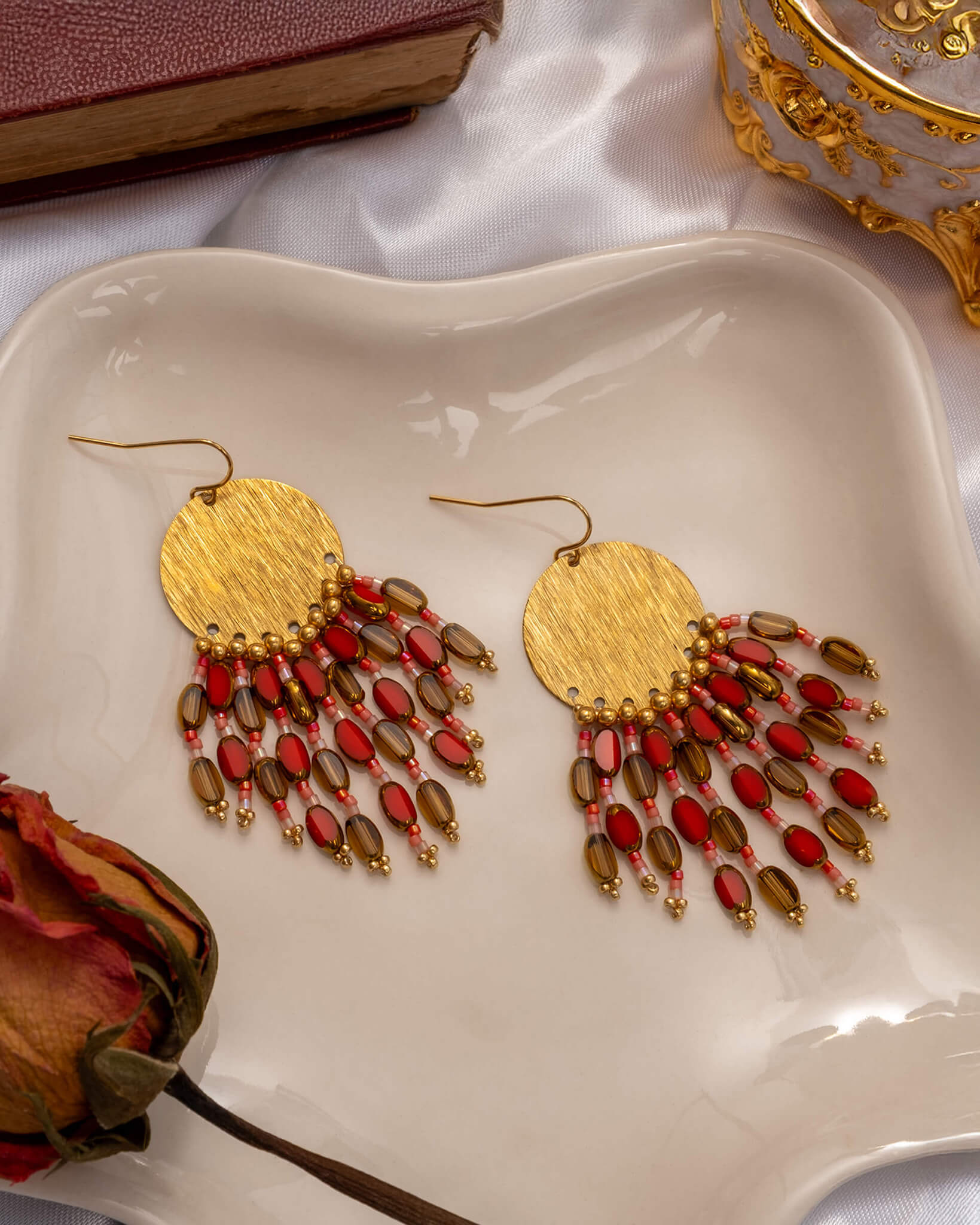 Gold earrings with red and brown beads on a white dish with a book and decorative box in the background.