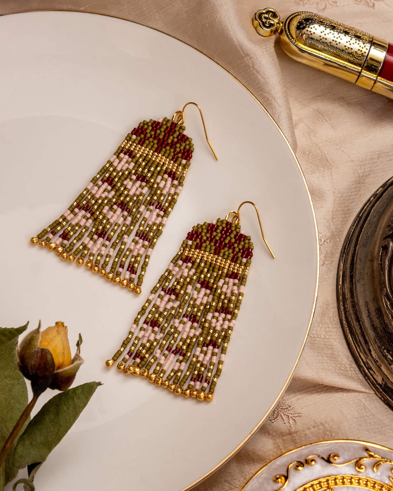 Pair of beaded earrings on a decorative plate with a rose and perfume bottle in the background.